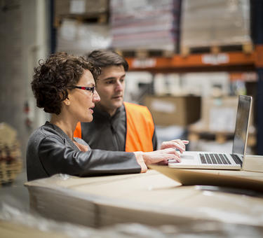 Two team members collaborate in a warehouse while looking at a laptop